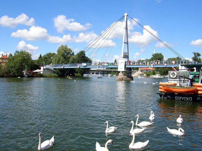 Weiße Schrägseilbrücke über einen See in Nikolaiken mit weißen Schwänen im Wasser und orangefarbenen Booten am Ufer unter blauem Himmel.