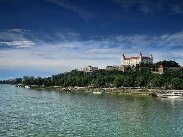 Die Burg Bratislava thront auf einem Hügel über der Donau mit Booten am Ufer und modernen Gebäuden im Hintergrund.