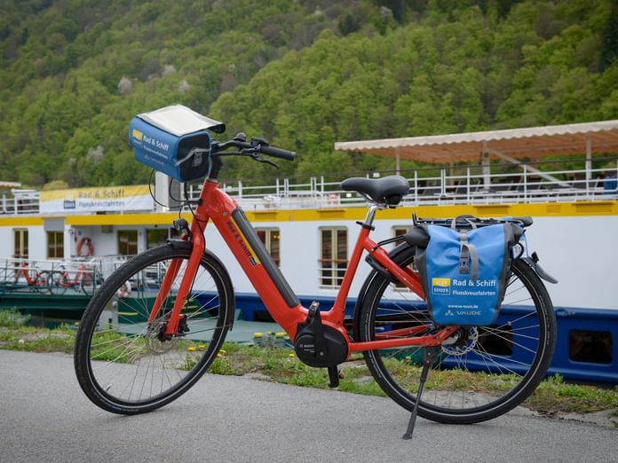 Rotes E-Bike mit blauen Packtaschen vor dem weißen Passagierschiff Prinzessin Katharina an der Donau, umgeben von grünen Hügeln.