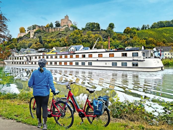 Cyclist with red bike watching white river cruise ship passing by. Castle on hilltop with vineyards visible in background under blue sky.