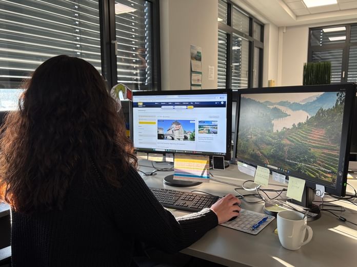 Frau mit dunklen Haaren sitzt am Büroschreibtisch und arbeitet an zwei Monitoren, einer zeigt eine Reise-Website, der andere ein Landschaftsbild.