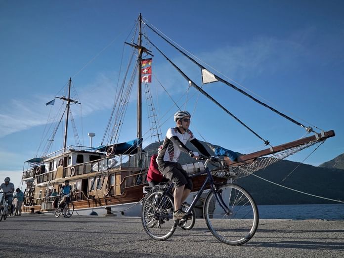 Radfahrer mit Helm auf Fahrrad vor traditionellem Holzsegelschiff Panagiota im Hafen, mit Bergen im Hintergrund.