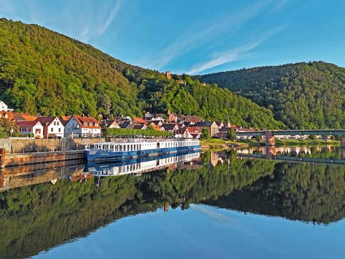 River cruise ship Casanova moored along a riverside village with colorful houses, forested hills, and perfect water reflections under blue sky.