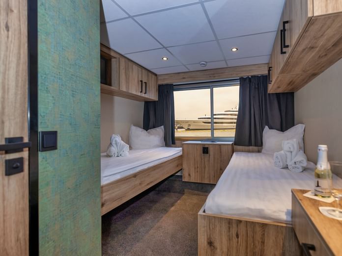Moderne Oberdeckkabine auf MS PRINCESS mit zwei Einzelbetten, Holzmöbeln, großem Fenster und blauen Vorhängen. Weiße Handtücher auf Betten.