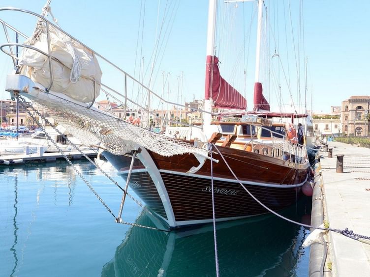 Traditionelles Holzsegelschiff Sundial mit dunklem Rumpf und roten Segeln in einem Mittelmeerhafen mit türkisfarbenem Wasser und Gebäuden.