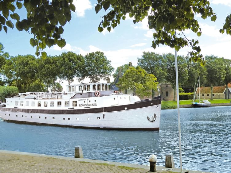 Weißes Passagierschiff Floor van Aemstel mit dunklem Rumpf auf Wasserstraße, eingerahmt von Baumzweigen. Historische Gebäude im Hintergrund.