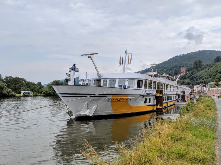 MS Classica river cruise ship with white and yellow hull moored along a green riverbank. Hills and buildings visible in background under cloudy sky.
