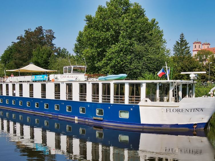 MS Florentina, ein blau-weißes Flusskreuzfahrtschiff mit mehreren Decks und großen Fenstern, vor Anker auf ruhigem Wasser mit Bäumen und Gebäuden im Hintergrund.