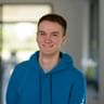 Fabian Homann in blue hoodie smiling Portrait of Fabian Homann wearing a blue hoodie, smiling warmly at the camera in a modern office setting with windows in the background.