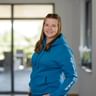 Ricarda Müller wearing a blue hoodie with white text on the sleeve, smiling at the camera in a modern office building with large windows.